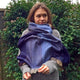 A person wrapped in a large, blue scarf stands in a garden with a wooden fence and leafy green plants in the background.