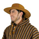 A person is smiling, wearing a brown wide-brimmed hat and a striped poncho with brown, yellow, and white patterns, against a plain white background.