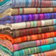 Wholesale lot of 15 queen alpaca wool blankets – colorful striped woolen blankets stacked neatly, displaying a range of vibrant hues, including blues, purples, oranges, and browns, creating a warm and cozy assortment in a retail setting