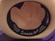 A Panama hat with a black inner band displays text: Ecuador Hats, Genuine Panama, Handwoven in Ecuador, CA#00076, 100% Paja Toquilla. The hat rests on a purple surface.