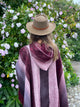 A person wearing a brown hat and a striped maroon shawl stands facing vibrant green bushes with light purple flowers, on a cobblestone path.