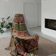 A patterned blanket drapes over a modern gray chair, situated on a beige rug in a minimalist room with a fireplace and a potted plant nearby.