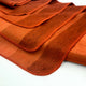 Cusubamba baby alpaca wool blanket – orange fabric lies in wavy folds, showcasing its stitched edges and gradient hues, on a smooth, light-colored surface