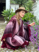 A person wearing a brown hat and a patterned shawl crouches beside purple flowers on a cobblestone path, surrounded by lush green plants and a white building facade.