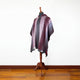 A maroon and gray striped poncho with a hood is displayed on a wooden mannequin. It has a button at the neckline and fringe hem, set against a plain white wall with a wooden floor.
