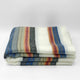Siguitag alpaca wool throw blanket – a folded blanket displays multicolored stripes, including blue, gray, and rusty red, against a white background, lying on a smooth, white surface