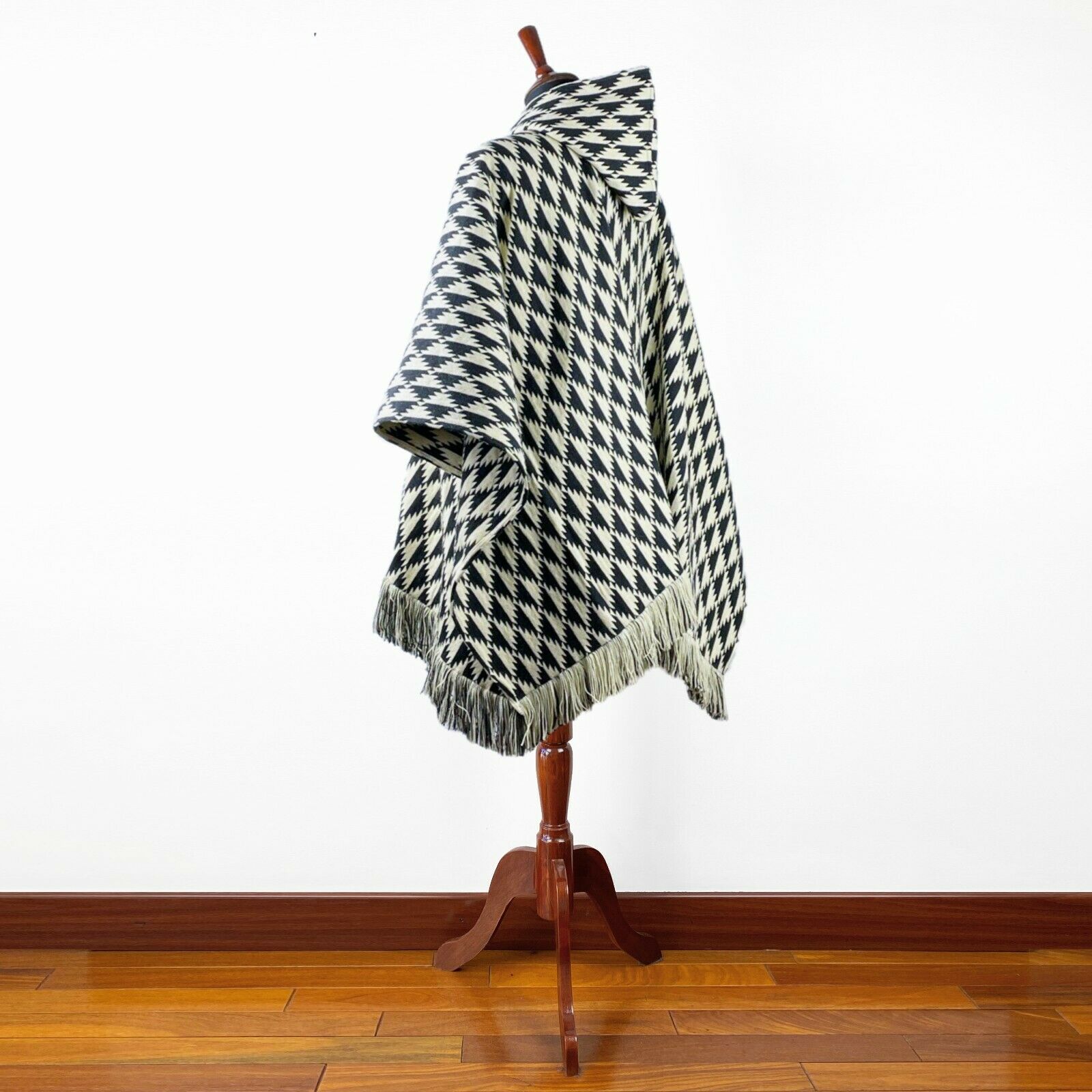 A mannequin displays a black and white houndstooth-patterned poncho with fringed edges, standing on a polished wooden floor against a plain white wall.