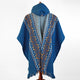 A blue hooded poncho features intricate geometric patterns in orange and white, displayed on a wooden stand against a plain white background.