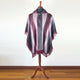 A maroon and gray poncho with fringe detailing hangs on a wooden mannequin stand. The poncho features a button closure and is displayed in a room with wooden flooring and a plain wall.