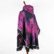 A multicolored poncho features a hood and black fringe edges, displayed on a wooden mannequin stand. The poncho's tie-dye pattern blends pink and purple hues, set against a plain white background.