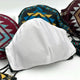 A white cloth face mask rests among colorful masks with geometric patterns, positioned on a light surface. The scene emphasizes the contrast between the plain and patterned fabrics.