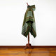 A green hooded poncho with white stripes drapes over a wooden mannequin stand, placed on a polished wooden floor against a plain white wall.