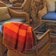 Yacupungu baby alpaca wool blanket – a striped orange and red blanket drapes over a wicker chair. Nearby, blue-cushioned wicker chairs surround a glass table, set in a cozy, rustic room with stone walls