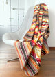 A patterned blanket, draped over a modern grey chair, features geometric and striped designs in warm colors. The setting is an indoor room with wooden flooring and frosted glass walls.