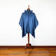 A blue hooded cape displayed on a wooden mannequin stand, set against a plain white wall and wooden floor.