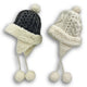 Two knit hats feature pom-poms, earflaps, and braided ties. One is black, the other white. Both have faux fur lining and trim, displayed on a plain white background.