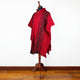 A red poncho with black geometric patterns is displayed on a wooden mannequin stand in a room with a wooden floor and white walls.