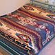 A bed is covered with a thick blanket featuring intricate geometric patterns in red, blue, and cream colors. The bed is in a room with light walls and carpet flooring.