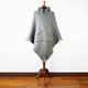A gray, hooded poncho with a front pocket is displayed on a wooden mannequin stand. It is set against a plain white wall and wooden floor background.