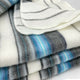 Teinza baby alpaca wool blanket – a neatly folded, soft blanket displays alternating stripes of grey, blue, and white, resting on a smooth white surface, suggesting a cozy, inviting atmosphere