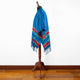 A vibrant blue poncho with colorful fringe and geometric patterns is draped over a wooden mannequin stand, set against a plain white wall and a wooden floor.