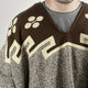 A person is wearing a patterned, brown and cream poncho with geometric and circular designs on the top section. The background is plain, emphasizing the textile details.