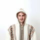 A person wears a hooded, knitted sweater with geometric patterns in earthy tones, standing against a plain white background.