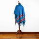 A blue poncho drapes on a wooden mannequin, with vibrant multicolored patterns and fringes at the bottom, set against a plain white wall and wooden floor.