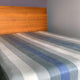 Poguios baby alpaca wool blanket – wooden headboard stands against a gray wall, with a bed covered by a striped blanket in shades of blue, gray, and beige, suggesting a minimalist bedroom setting