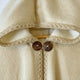 A beige woolen garment features an embroidered hood and two large brown buttons, hanging from a light wooden rod in a neutral indoor setting.