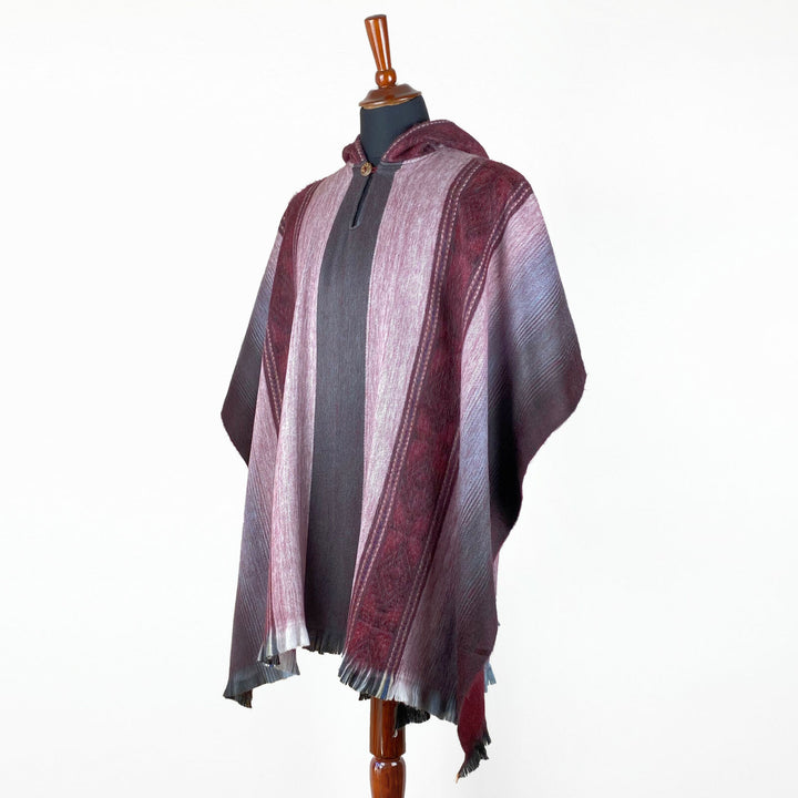 A mannequin displays a hooded, striped poncho with shades of burgundy and gray, featuring a button closure and fringed hem, set against a plain white background.