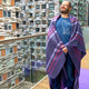 Punguloma baby alpaca wool blanket – a person stands wrapped in a purple plaid blanket, wearing a shirt with a line-drawn animal and text: save the alpacas. They are on a balcony with modern apartment buildings visible behind