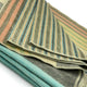 Cojitambo alpaca wool throw blanket – striped blankets are neatly stacked, highlighting alternating colors of green, blue, orange, and beige in a soft woolen fabric, against a plain white background