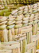 Woven basket features tightly interlaced natural fibers with varying shades of beige and green, detailed with small brown spots. It sits among similar baskets, emphasizing craftsmanship and texture.