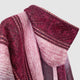 A folded, woolen fabric displays rich maroon and pink colors with intricate patterns, draped against a plain background. The texture appears soft and the design is detailed and woven.