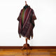 A mannequin wearing a multicolored, intricately patterned poncho is displayed on a wooden stand against a plain white background, with a polished wooden floor beneath.