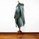 A green, textured poncho drapes over a wooden mannequin stand, set in a room with a polished wooden floor and plain white wall.