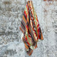 A colorful, patterned blanket hangs against a weathered, textured wall. The design includes geometric shapes in red, orange, green, and brown hues, creating a warm and vibrant appearance.