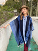 A person in a blue poncho and brown hat is standing, hand on a white wall, surrounded by a leafy, outdoor setting.