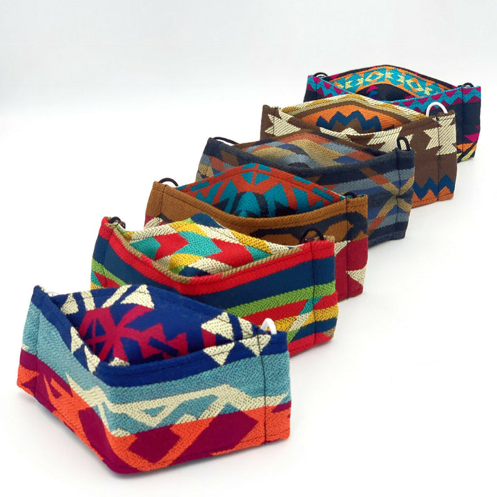 Six colorful, patterned fabric face masks are lined up diagonally against a plain white background, each featuring various geometric designs and vibrant hues including blue, red, and orange.