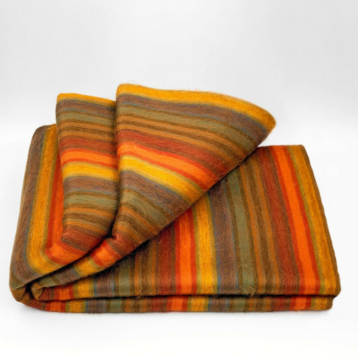 Cumanda baby alpaca wool blanket – a folded woolen blanket displays vibrant, multicolored horizontal stripes in orange, yellow, and brown hues, resting against a plain white background