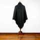 A black hooded poncho with fringed edges is draped over a wooden stand, set against a plain white wall and wood floor background.