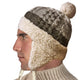 A man wears a knitted winter hat with ear flaps and a pom-pom, featuring a textured wool pattern. He faces left against a plain white background, wearing a beige sweater.
