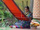 A woman weaves on a bright orange loom, seated on a patio with lush plants around her and a brick wall in the background. Text on the image reads BVelarde.