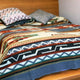 A bed is covered with a blanket featuring geometric patterns in earth tones, accompanied by two patterned pillows, in a room with a wooden headboard and plain wall.