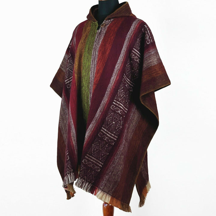 A colorful woven poncho with geometric patterns drapes over a mannequin. The poncho features maroon, red, and green vertical stripes in a minimalist studio setting.