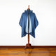 A dark blue cape with a hood is displayed on a wooden mannequin stand, situated in a room with white walls and a wooden floor.