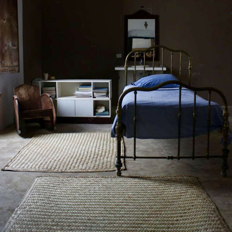 A brass bed with blue bedding stands in a sparsely furnished bedroom. Nearby, a wooden chair and a shelf filled with books rest against dark walls, complemented by two woven rugs.