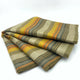 Rumi baby alpaca wool blanket – a folded striped fabric with yellow, orange, and gray hues lies on a plain white surface, showcasing its soft texture and color pattern in a neatly arranged stack