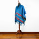 A vibrant blue poncho with colorful patterns hangs on a wooden mannequin stand. The poncho has fringed edges and is displayed against a plain white wall and wooden floor.